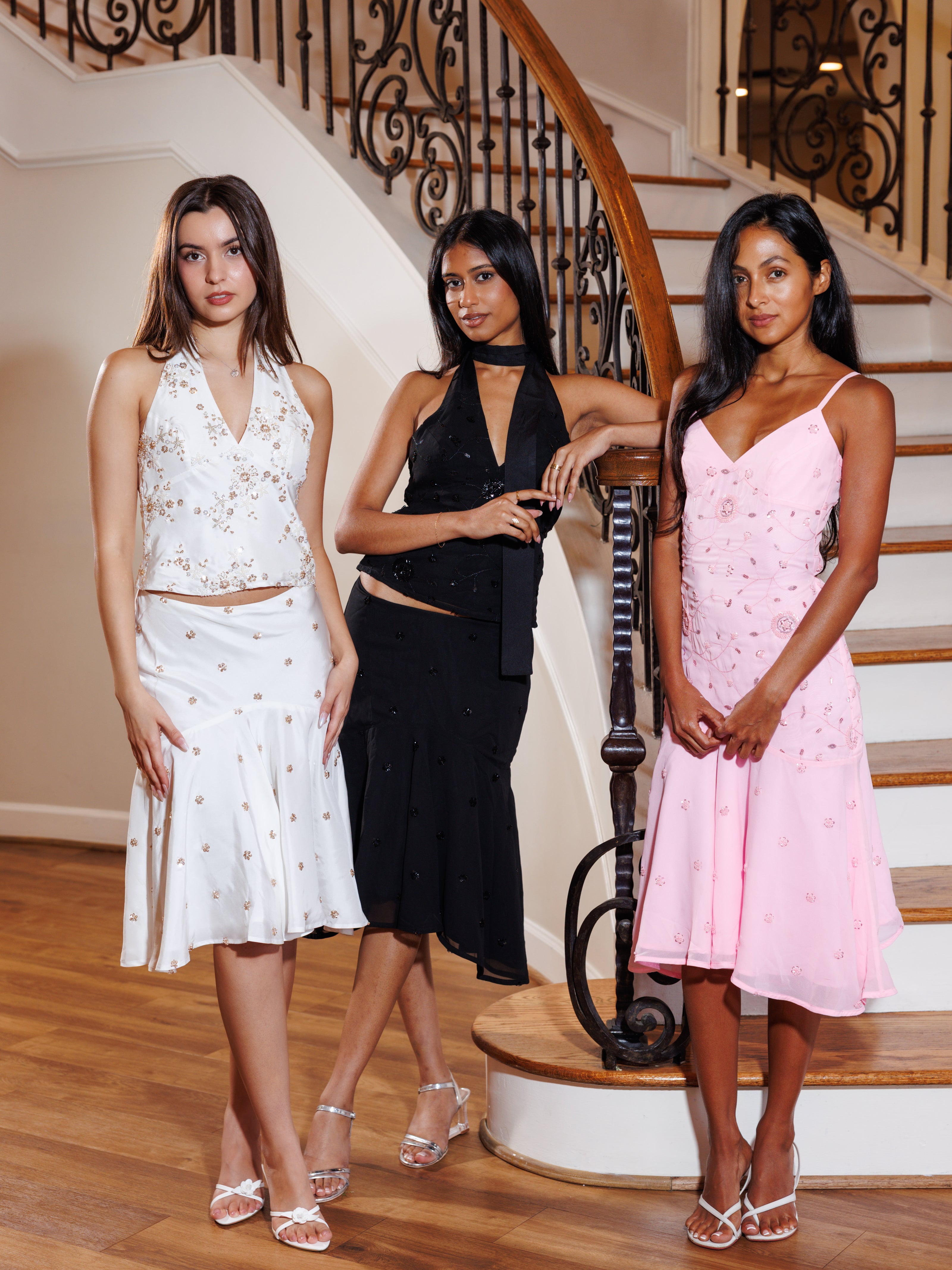 Three women posing together on a wooden staircase indoors, wearing elegant dresses: a white sleeveless crop top with a matching skirt, a black halter-neck dress, and a light pink spaghetti-strap dress with subtle floral embroidery.
