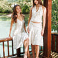 Two women modeling matching white sleeveless dresses with gold floral embroidery on a wooden deck beside a body of water, with trees in the background.
