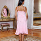 A woman stands with her back to the camera wearing a light pink sleeveless midi dress with thin straps and a ruffled skirt, posed in an elegant room with a large mirror, vanity table, and floral rug.