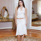 A woman stands in a vintage-style room wearing a white sleeveless halter crop top and matching white skirt with small gold floral details. She is posed in front of a large ornate mirror and a gold console table.