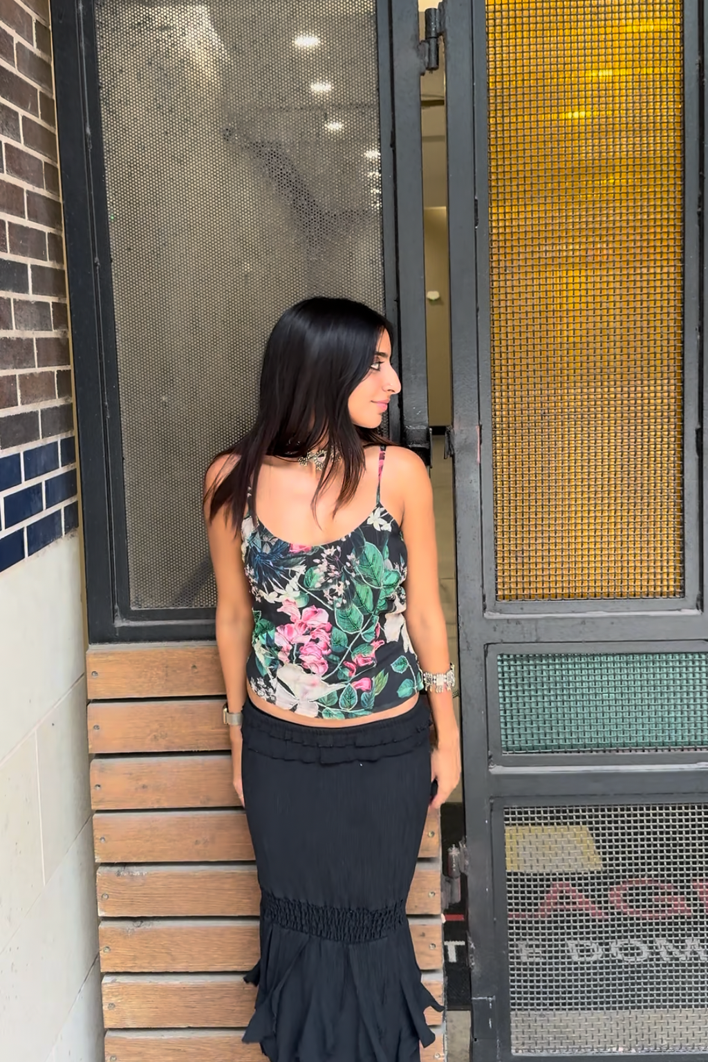 Woman in floral tank top and black skirt standing against a building entrance.